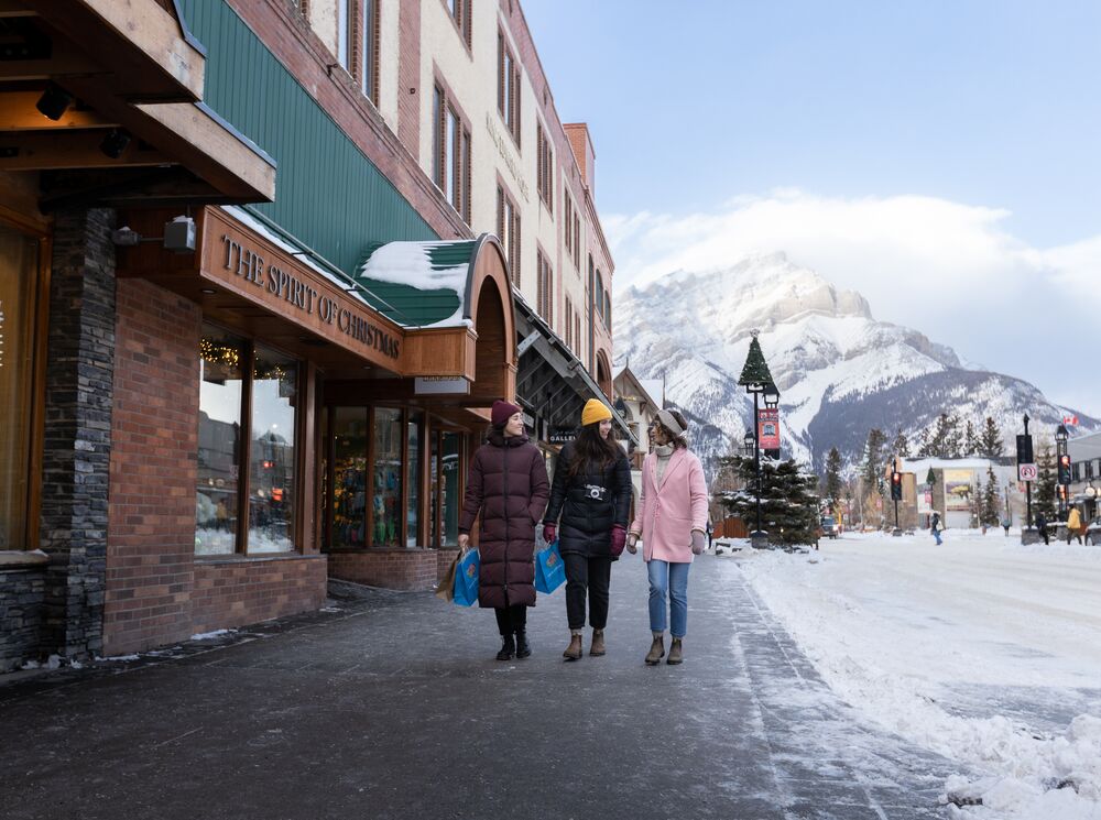 Three girls walk down Banff Ave while shopping with Cascade Mountain in the background.